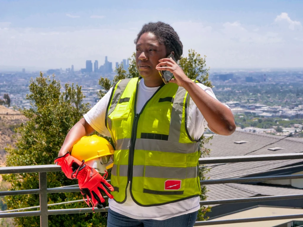 Black woman in safety vest and hardhat holding phone with cityscape background.